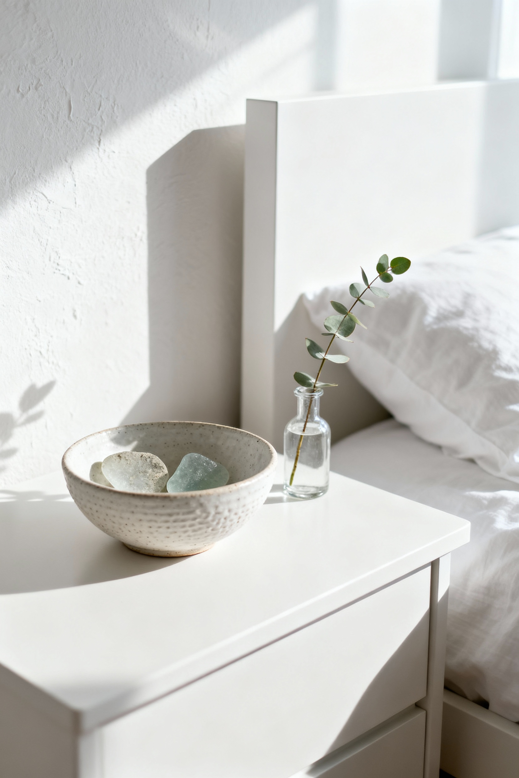 A close-up of a beautifully curated bedside vignette in a white bedroom, featuring a ceramic bowl, sea glass, and eucalyptus, embodying serene personal touches.