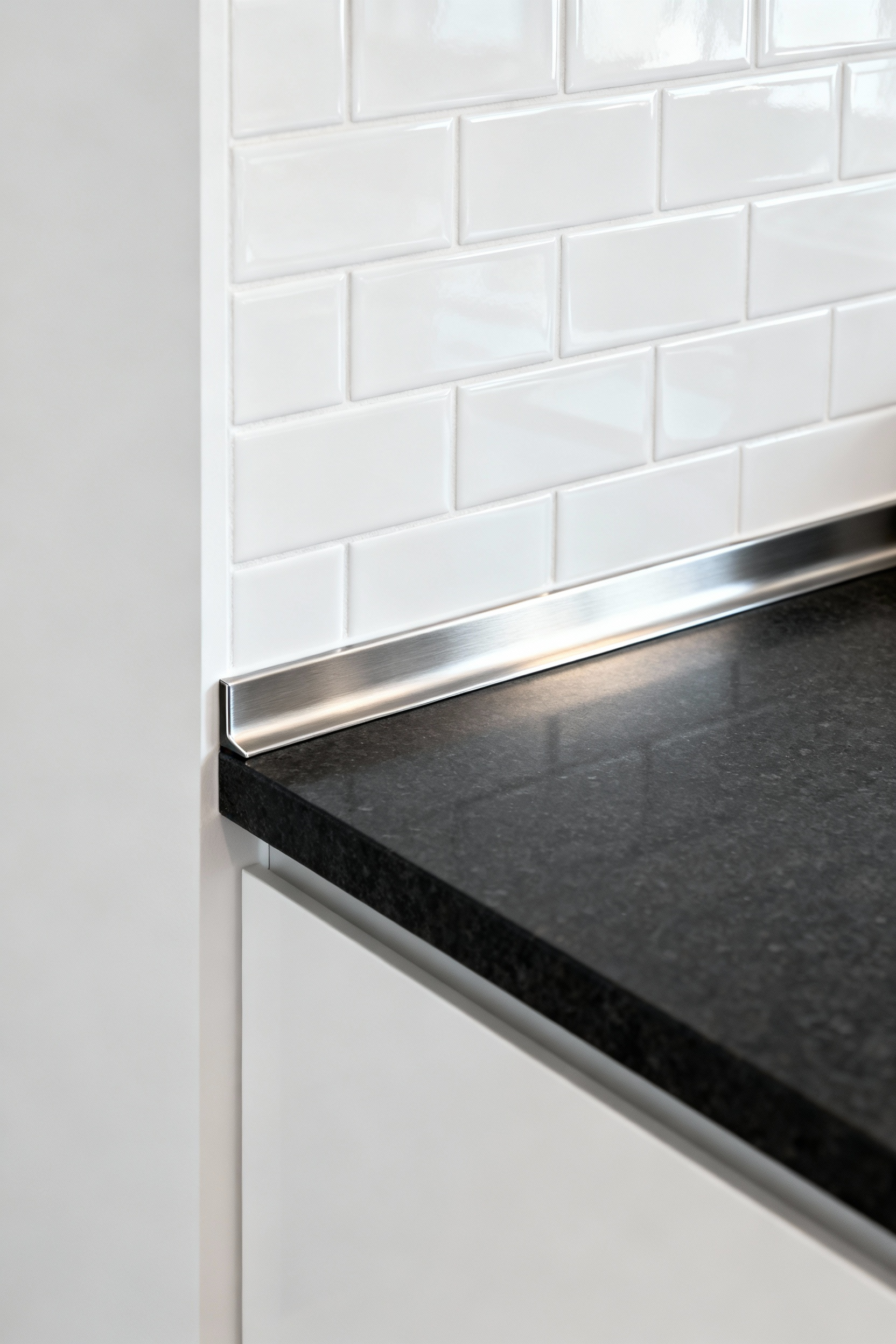 Close-up of a kitchen backsplash featuring a professional, seamless brushed stainless steel L-angle edging profile at the transition to a dark quartz countertop in a modern urban interior.