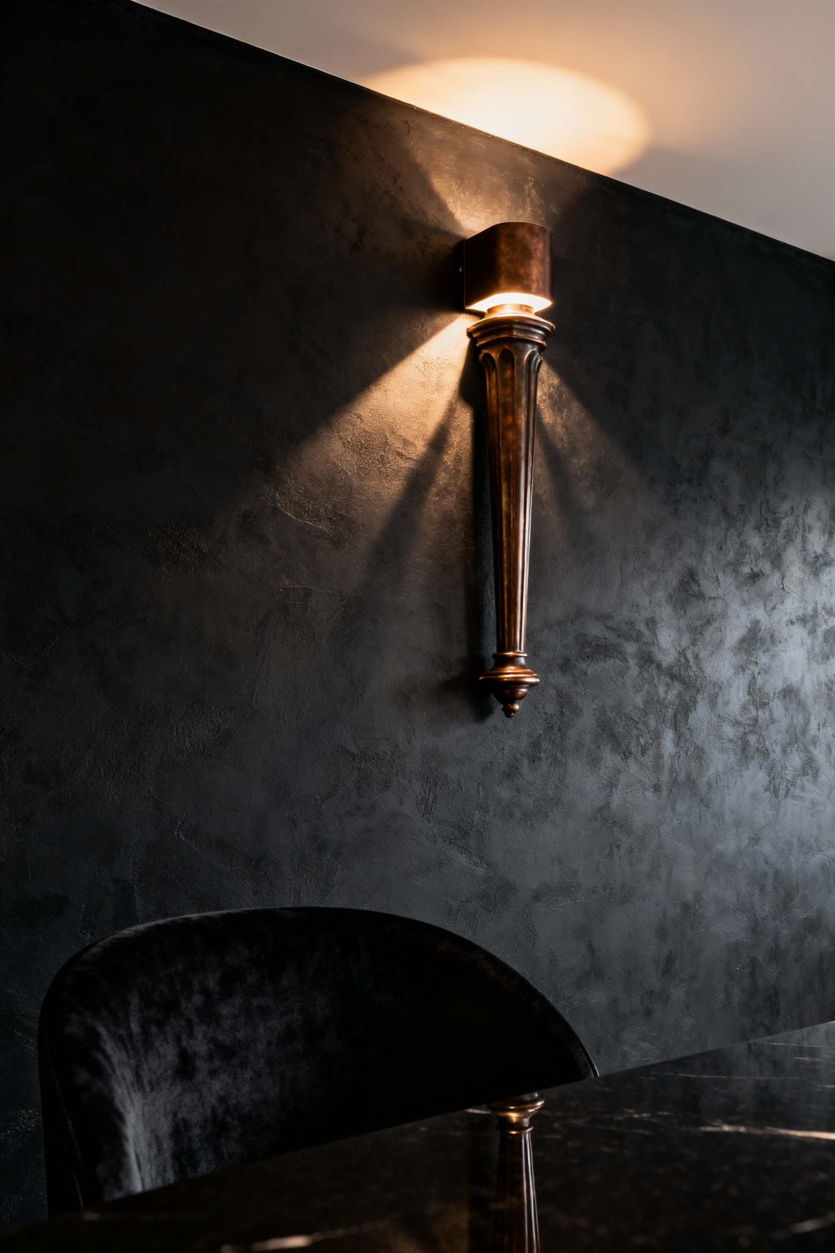 A luxurious black dining room featuring a sculptural bronze architectural sconce on a matte black plaster wall, casting a warm sidelight and dramatic shadows, enhancing the room's elegant and moody atmosphere.