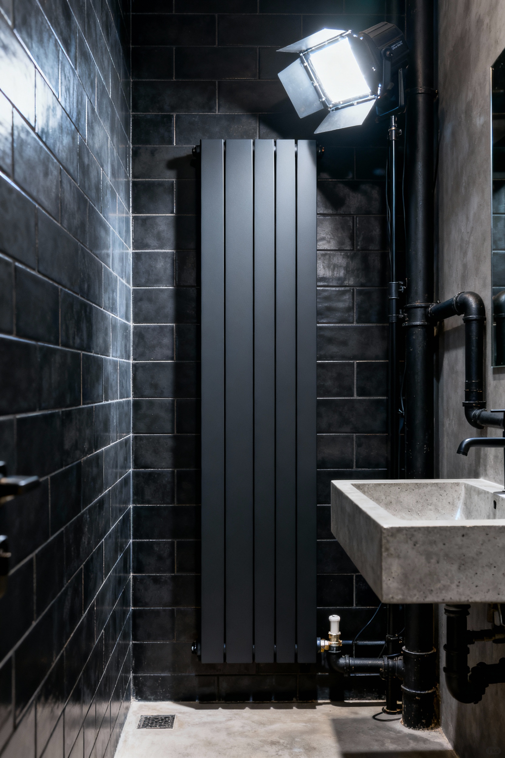Narrow matte black vertical radiator maximizing heating surface area in a small, industrially designed bathroom layout with dark tile and polished concrete fixtures.